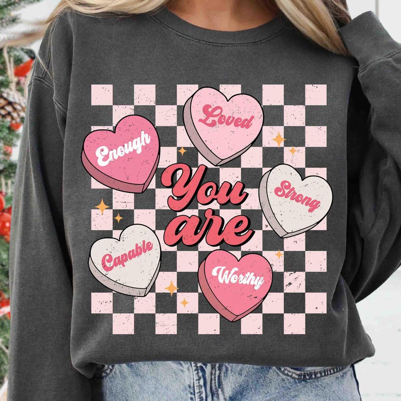 May include: A charcoal gray sweatshirt with a checkered pattern and pink hearts that say "Loved", "Enough", "Strong", "Capable", and "Worthy". The text "You are" is written in a retro font.
