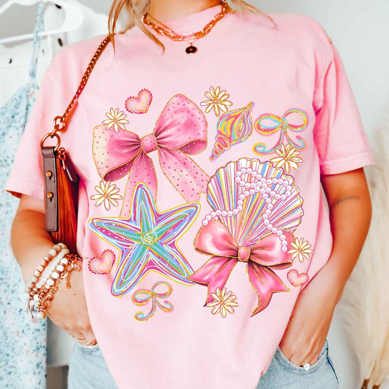 Op de afbeelding: Zachtroze T-shirt met een grafisch ontwerp van een zeester, schelpen, strikken en bloemen. Het ontwerp bevat een grote roze strik met gouden accenten, een veelkleurige zeester en een schelp met een parelketting.