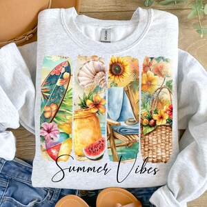May include: A gray sweatshirt with a watercolor design of a beach scene. The design includes a surfboard, a beach chair, a basket of fruit, and a watermelon. The text "Summer Vibes" is printed below the design.