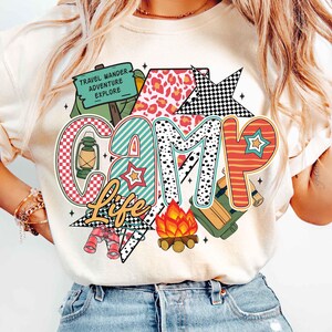 Puede incluir: Una camiseta blanca con un dise&ntilde;o gr&aacute;fico colorido que dice "Camp Life". El dise&ntilde;o incluye una fogata, una linterna, binoculares, una mochila y una estrella con un patr&oacute;n a cuadros. Las palabras "Travel Wander Adventure Explore" est&aacute;n en un letrero en el dise&ntilde;o.