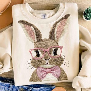 May include: A cream-colored sweatshirt with an embroidered image of a brown bunny wearing pink glasses and a pink bow tie.