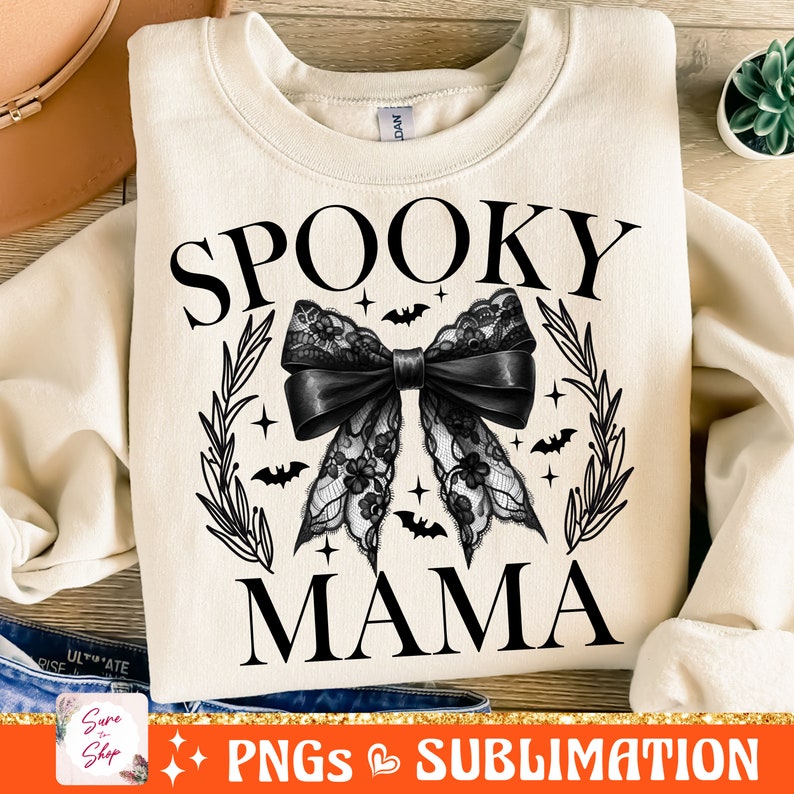 May include: A cream-colored sweatshirt with the text "Spooky Mama" in black. A black lace bow with bats and stars is centered on the sweatshirt.