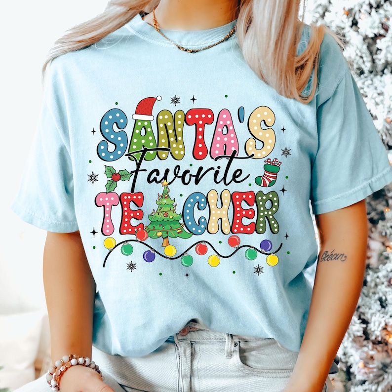 May include: A light blue t-shirt with a colorful graphic that says "Santa's Favorite Teacher" with a Christmas tree and lights.