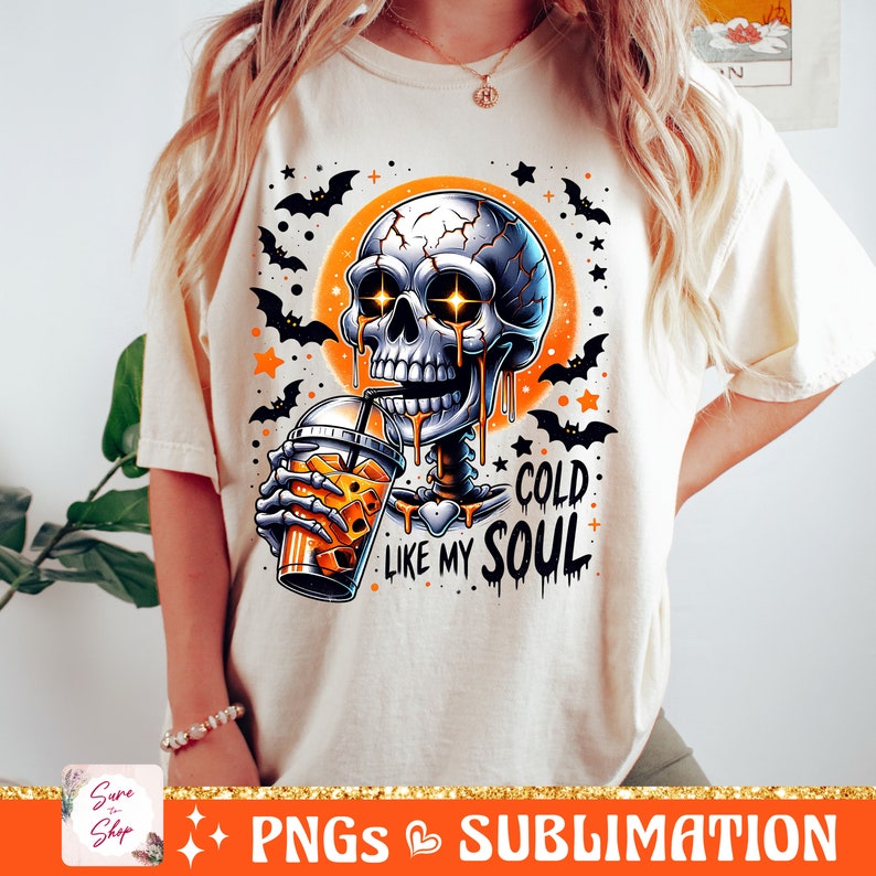 May include: A white t-shirt with a graphic of a skeleton drinking a beverage with a skull face and glowing eyes. The text "Cold Like My Soul" is printed below the skeleton. The background is orange and black with bats and stars.