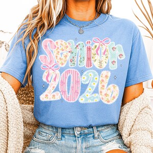 Op de afbeelding: Lichtblauw T-shirt met de tekst "Senior 2026". Het woord "Senior" is in een decoratief lettertype met strikken, en de cijfers "2026" zijn in een vergelijkbare stijl met madeliefjes en strepen.