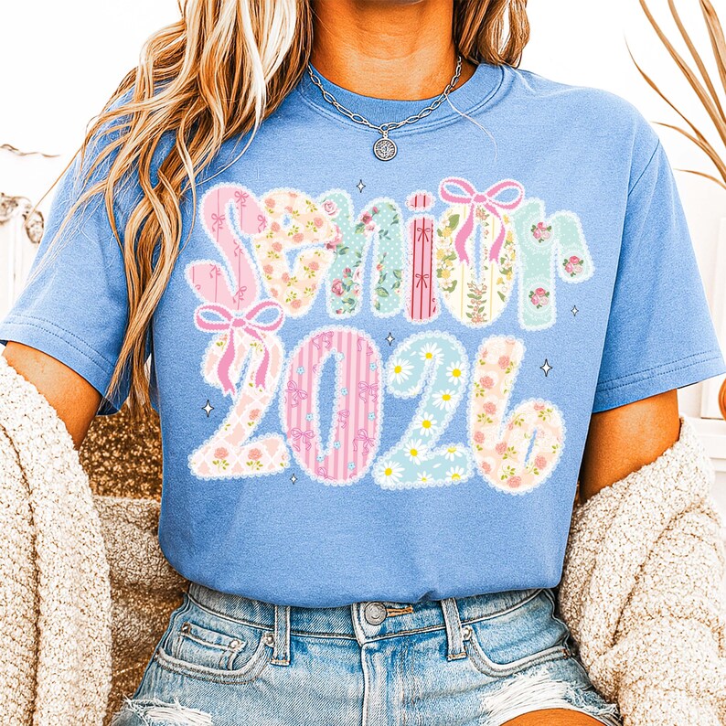 Op de afbeelding: Lichtblauw T-shirt met de tekst "Senior 2026". Het woord "Senior" is in een decoratief lettertype met strikken, en de cijfers "2026" zijn in een vergelijkbare stijl met madeliefjes en strepen.
