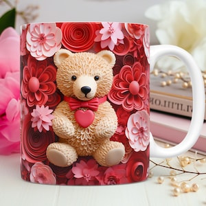 May include: White ceramic mug featuring a teddy bear design with a red bow tie and heart. The mug is decorated with red and pink floral accents. The handle is white.