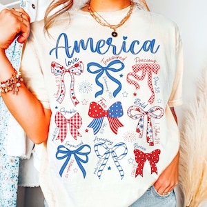 Puede incluir: Camiseta color crema con la palabra "America" en escritura azul. La camiseta presenta varios dise&ntilde;os de lazo en rojo, blanco y azul, con palabras como "love", "free" y "brave" incorporadas en el dise&ntilde;o. La camiseta tiene un tema patri&oacute;tico informal.