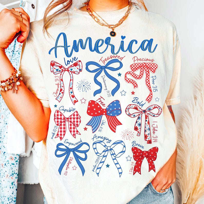 Puede incluir: Camiseta color crema con la palabra "America" en escritura azul. La camiseta presenta varios dise&ntilde;os de lazo en rojo, blanco y azul, con palabras como "love", "free" y "brave" incorporadas en el dise&ntilde;o. La camiseta tiene un tema patri&oacute;tico informal.