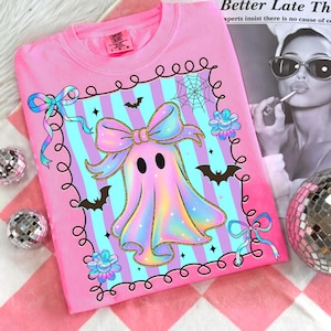 Puede incluir: Sudadera rosa Comfort Colors con un dise&ntilde;o de fantasma pastel, un gran lazo, murci&eacute;lagos y una telara&ntilde;a. El fantasma est&aacute; sobre un fondo a rayas azul y morado. La camisa tiene cuello redondo y una peque&ntilde;a etiqueta en la parte superior izquierda.