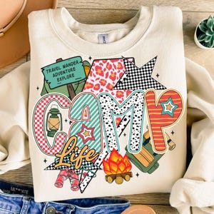 Puede incluir: Una sudadera beige con un dise&ntilde;o gr&aacute;fico colorido que dice "Camp Life". El gr&aacute;fico incluye una tienda de campa&ntilde;a, una fogata, una linterna, binoculares y una estrella. El texto "Travel Wander Adventure Explore" est&aacute; en un letrero verde.