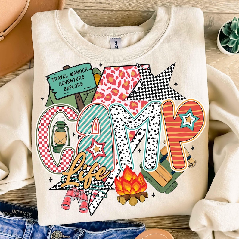 Puede incluir: Una sudadera beige con un dise&ntilde;o gr&aacute;fico colorido que dice "Camp Life". El gr&aacute;fico incluye una tienda de campa&ntilde;a, una fogata, una linterna, binoculares y una estrella. El texto "Travel Wander Adventure Explore" est&aacute; en un letrero verde.