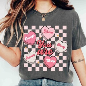 May include: A gray t-shirt with a checkered pattern and pink hearts that say "You are" with the words "Loved", "Enough", "Strong", "Capable", and "Worthy" on the hearts.