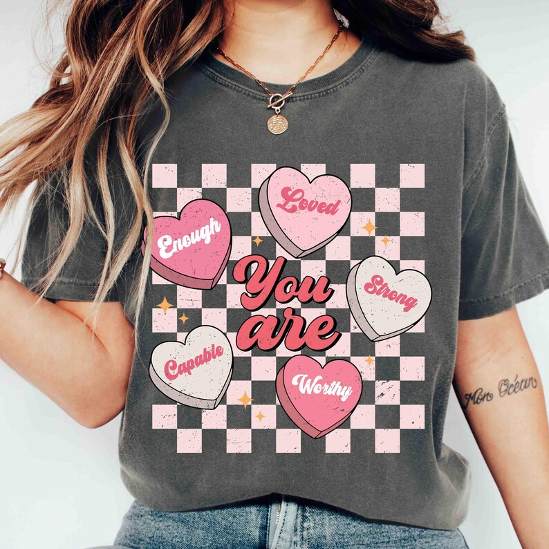 May include: A gray t-shirt with a checkered pattern and pink hearts that say "You are" with the words "Loved", "Enough", "Strong", "Capable", and "Worthy" on the hearts.