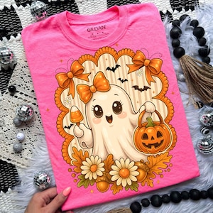 Pode incluir: Camiseta rosa com um desenho de fantasma de desenho animado segurando um doce e uma ab&oacute;bora. O fantasma tem um la&ccedil;o laranja e &eacute; cercado por decora&ccedil;&otilde;es de outono. Gola redonda, mangas curtas.
