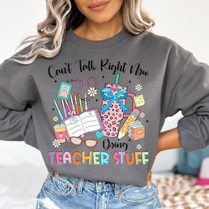 May include: Gray sweatshirt with a colorful graphic design that says "Can't Talk Right Now Doing Teacher Stuff". The graphic includes a coffee cup, books, pencils, and glasses.