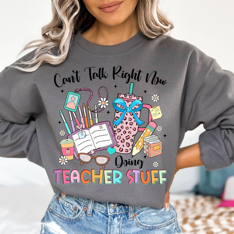 May include: Gray sweatshirt with a colorful graphic design that says "Can't Talk Right Now Doing Teacher Stuff". The graphic includes a coffee cup, books, pencils, and glasses.