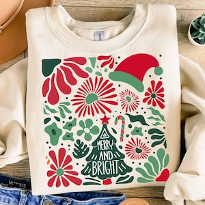 Puede incluir: Sudadera de color crema con un dise&ntilde;o navide&ntilde;o festivo. El dise&ntilde;o presenta elementos florales rojos y verdes, un gorro de Pap&aacute; Noel, un bast&oacute;n de caramelo y un &aacute;rbol de Navidad con las palabras "Merry and Bright".
