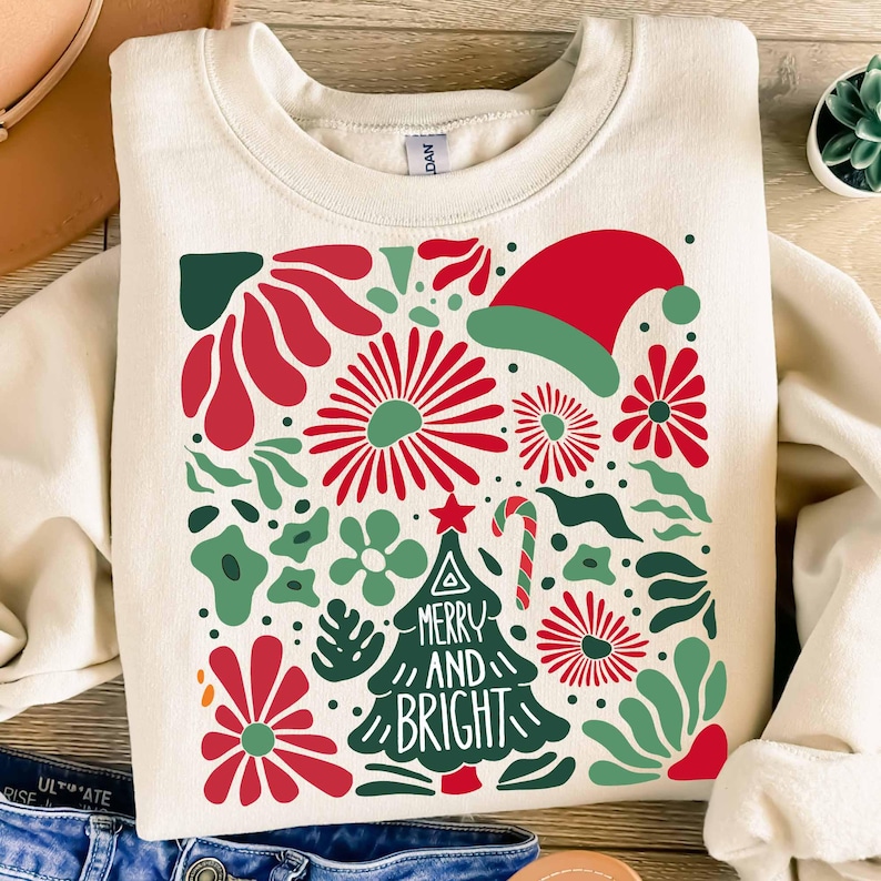 Puede incluir: Sudadera de color crema con un dise&ntilde;o navide&ntilde;o festivo. El dise&ntilde;o presenta elementos florales rojos y verdes, un gorro de Pap&aacute; Noel, un bast&oacute;n de caramelo y un &aacute;rbol de Navidad con las palabras "Merry and Bright".