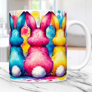 May include: White ceramic mug with a colorful watercolor design of four bunny rabbits with their backs turned to the viewer. The bunnies are blue, pink, yellow, and white.