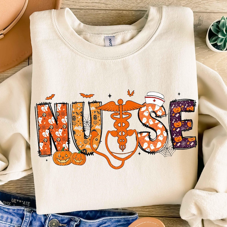 May include: Cream-colored sweatshirt with the word "NURSE" in orange, purple, and white Halloween-themed lettering. The design includes pumpkins, ghosts, bats, and a medical caduceus. A nurse's cap is also featured.