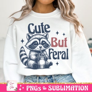 May include: White sweatshirt featuring a raccoon illustration drinking from a cup with a straw. The text reads "Cute But Feral" in navy and red. The design includes star accents.