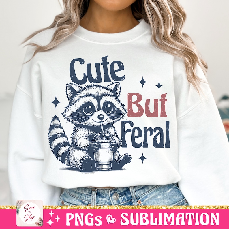 May include: White sweatshirt featuring a raccoon illustration drinking from a cup with a straw. The text reads "Cute But Feral" in navy and red. The design includes star accents.