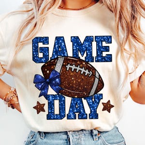 Puede incluir: Camiseta color crema con un gr&aacute;fico de f&uacute;tbol americano. Las palabras "GAME DAY" est&aacute;n en letras azules brillantes, con un bal&oacute;n de f&uacute;tbol marr&oacute;n y un lazo azul. Dos estrellas marrones flanquean la palabra "DAY".
