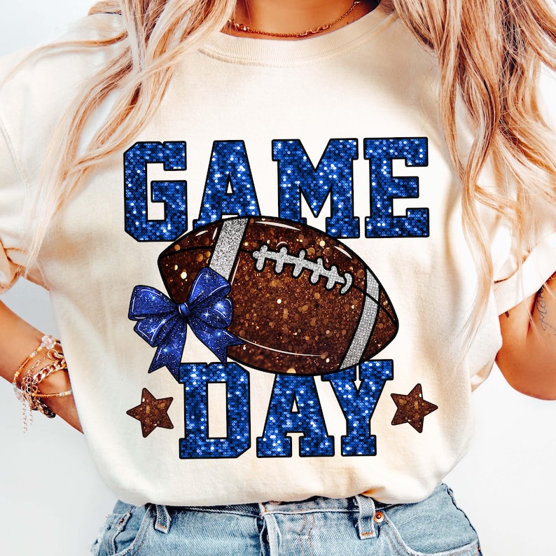 Puede incluir: Camiseta color crema con un gr&aacute;fico de f&uacute;tbol americano. Las palabras "GAME DAY" est&aacute;n en letras azules brillantes, con un bal&oacute;n de f&uacute;tbol marr&oacute;n y un lazo azul. Dos estrellas marrones flanquean la palabra "DAY".