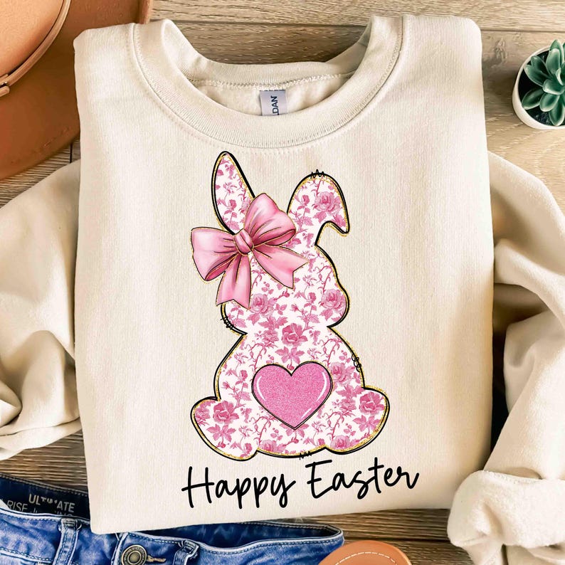 Op de afbeelding: Een cr&egrave;mekleurige trui met een roze bloemetjeskonijn met een roze strik en een roze hart. De tekst "Happy Easter" is onder het konijn gedrukt.