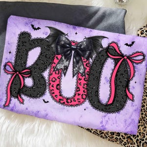 May include: A decorative pillow with the word "BOO" in black leopard print letters outlined with black lace. The "O" is pink leopard print. Black bat wings and bows adorn the design, with pink bows on the "B" and "O".