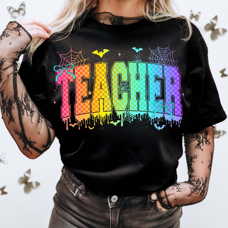 May include: Black t-shirt with the word "TEACHER" in rainbow checkered letters. The design includes spiderwebs, bats, and dripping paint, creating a Halloween theme. A bow is on the left side of the word.