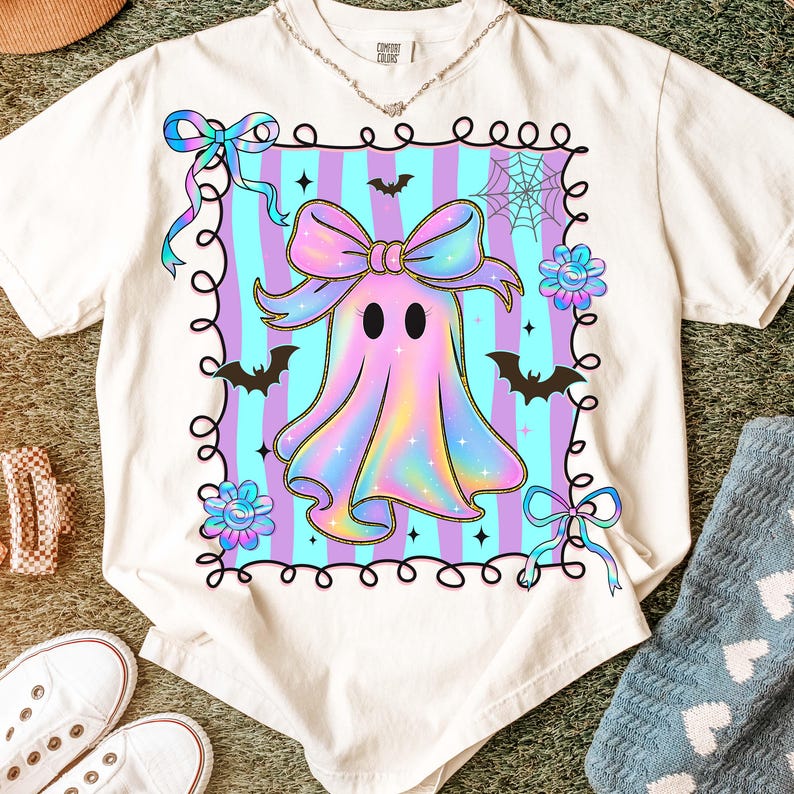 Puede incluir: Camiseta color crema con un dise&ntilde;o de fantasma pastel, un gran lazo, murci&eacute;lagos y flores. El fantasma est&aacute; rodeado por un marco decorativo con un fondo a rayas. Un dise&ntilde;o divertido y caprichoso para Halloween.