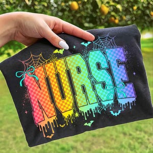 Pu&ograve; includere: T-shirt nera con la scritta "NURSE" a grandi lettere colorate a scacchi. Le lettere passano dall'arancione al blu, con ragnatele, pipistrelli e dettagli di vernice gocciolante. Un fiocco turchese &egrave; sul lato sinistro.