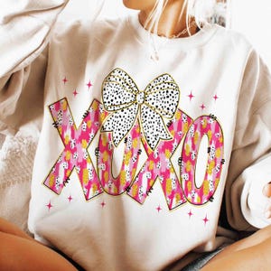 K&ouml;nnte beinhalten: Cremefarbenes Sweatshirt mit einer gro&szlig;en Schleife und den Buchstaben "XOXO" in einem rosa-goldenen Muster. Die Schleife ist wei&szlig; mit schwarzen Punkten. Rosa Sternakzente umgeben den Text.