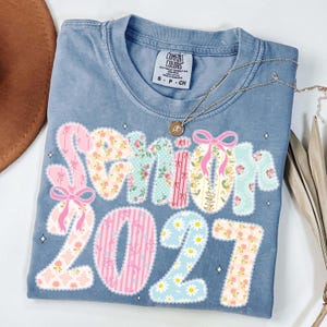 May include: A blue Comfort Colors t-shirt with the word "Senior" and the number "2021" in a floral patchwork design. The shirt has a crew neck and short sleeves. A gold necklace with a pendant is draped across the shirt.