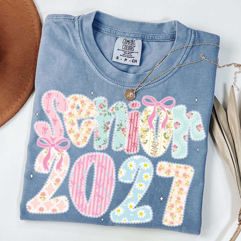 May include: A blue Comfort Colors t-shirt with the word "Senior" and the number "2021" in a floral patchwork design. The shirt has a crew neck and short sleeves. A gold necklace with a pendant is draped across the shirt.