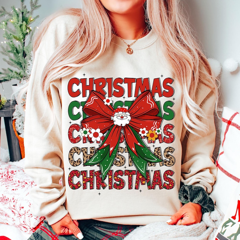 May include: A cream-colored sweatshirt with a festive Christmas design. The design features a red bow with a Santa face in the center, surrounded by green leaves and white flowers. The word "Christmas" is repeated in a stacked pattern, with each word having a different design element.