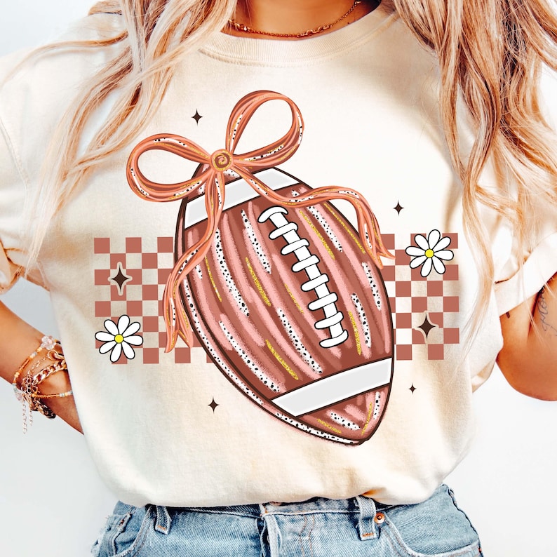 May include: Cream-colored t-shirt featuring a football design with a pink bow, white laces, and gold accents. The design includes a checkered pattern, daisies, and star accents, creating a sporty and feminine aesthetic.