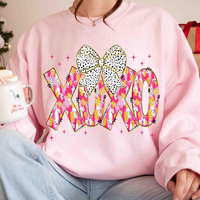 K&ouml;nnte beinhalten: Ein hellrosa Sweatshirt mit den Buchstaben "XOXO" in einem rosa-goldenen Muster und einer wei&szlig;en Schleife mit schwarzen Punkten. Das Design ist mit goldenem Glitzer und rosa Sternen akzentuiert.