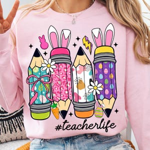 May include: Pink sweatshirt featuring a design of four colourful pencils with bunny ears and Easter egg patterns. The text '#teacherlife' is printed beneath the pencils.