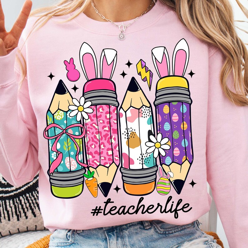 May include: Pink sweatshirt featuring a design of four colourful pencils with bunny ears and Easter egg patterns. The text '#teacherlife' is printed beneath the pencils.