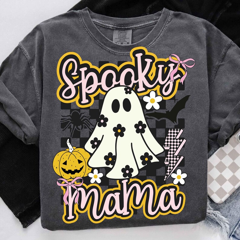 Puede incluir: Sudadera gris oscuro con un dise&ntilde;o de Halloween. El dise&ntilde;o presenta un fantasma con flores, una calabaza, una ara&ntilde;a, un murci&eacute;lago y las palabras "Spooky Mama" en una fuente retro. Los lazos rosas y los rayos completan el dise&ntilde;o.