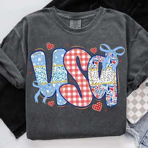 Puede incluir: Sudadera gris oscuro con la palabra "USA" en un dise&ntilde;o colorido. Las letras presentan patrones como estampado de leopardo, cuadros vichy e estampados florales, con lazos azules y detalles de coraz&oacute;n. Una prenda superior informal de tem&aacute;tica patri&oacute;tica.
