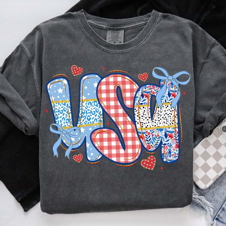 Puede incluir: Sudadera gris oscuro con la palabra "USA" en un dise&ntilde;o colorido. Las letras presentan patrones como estampado de leopardo, cuadros vichy e estampados florales, con lazos azules y detalles de coraz&oacute;n. Una prenda superior informal de tem&aacute;tica patri&oacute;tica.