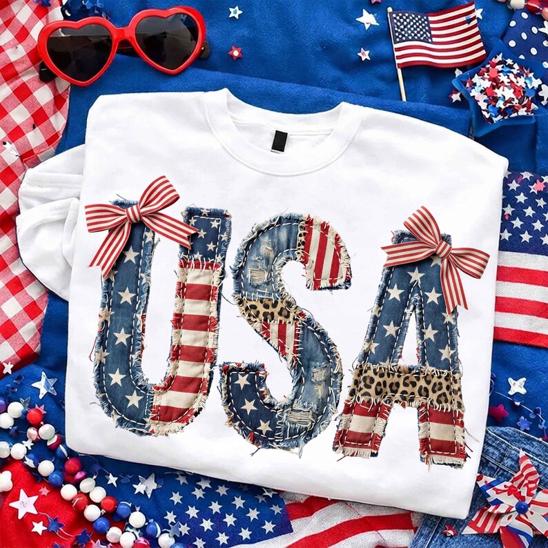 Op de afbeelding: Witte crewneck sweatshirt met het woord "USA" op de voorkant. De letters zijn gemaakt van denim, Amerikaanse vlagstof en luipaardprint. Elke letter heeft een rood-wit gestreepte strik. Patriotisch thema.