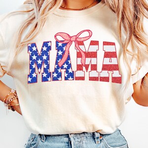 May include: A white t-shirt with a red, white, and blue "MAMA" graphic with a pink bow on top. The graphic is designed to look like the American flag.