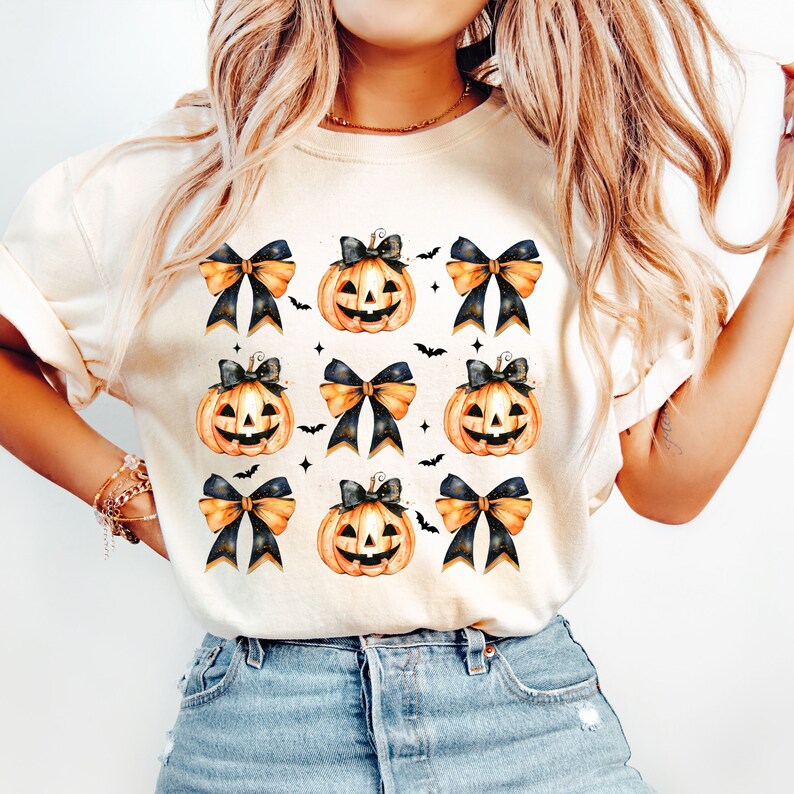 May include: A white t-shirt with a pattern of orange and black pumpkins with black bows and bats. The pumpkins have a watercolor effect.
