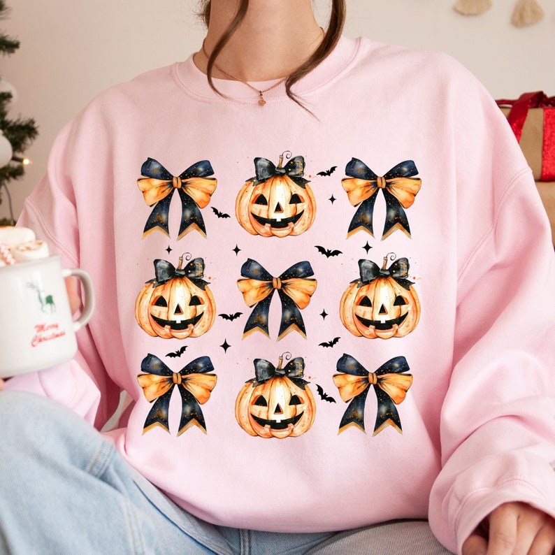 May include: A pink sweatshirt with a repeating pattern of orange pumpkins with black eyes and mouths, black and gold bows, and black bats.
