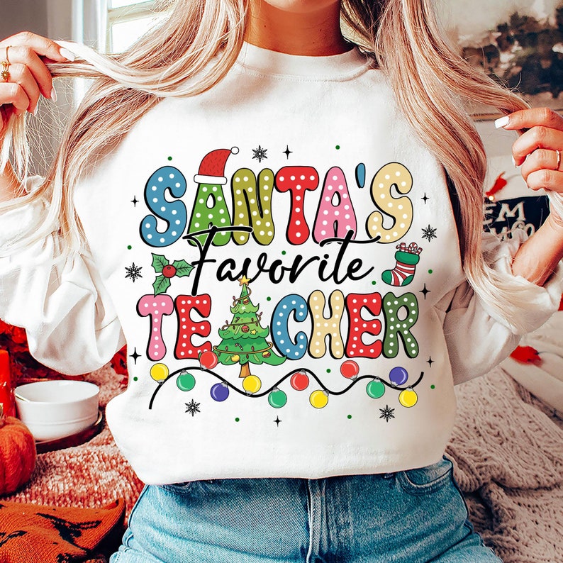 May include: White sweatshirt with a colorful graphic that says "Santa's Favorite Teacher" with a Christmas tree, ornaments, and a stocking.